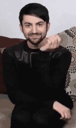 a man with a beard is sitting on a couch with his hands folded .