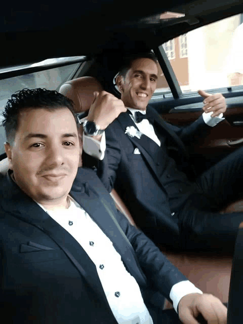 two men in suits are sitting in a car and one is giving a thumbs up