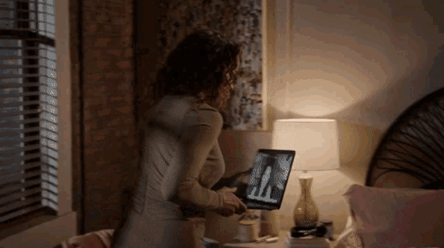 a woman is using a laptop in a bedroom with a lamp and a window