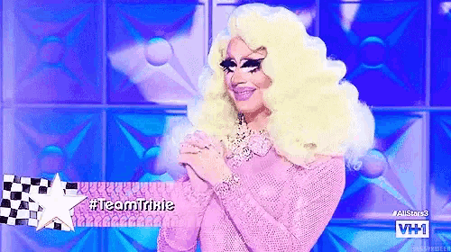 a drag queen is wearing a pink sweater and has the hashtag #teamtrixie on her face
