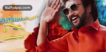 a man wearing sunglasses and a red shirt is smiling and waving his hand .