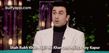 a man in a white suit and black turtleneck is talking to shah rukh khan