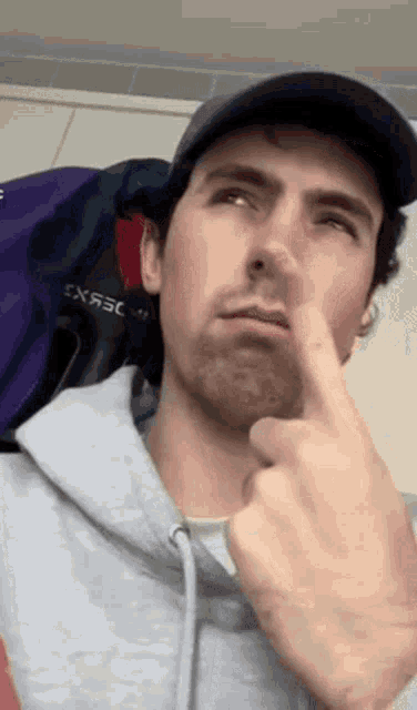 a man wearing a hat and a hoodie is pointing at his nose .