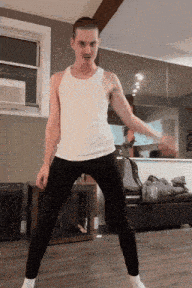 a man wearing a white tank top and black pants is dancing in a living room