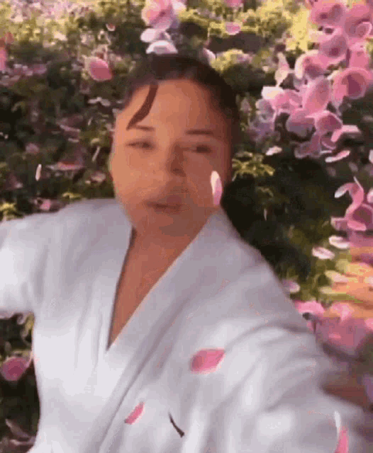 a woman in a white robe stands in front of a bush of pink flowers