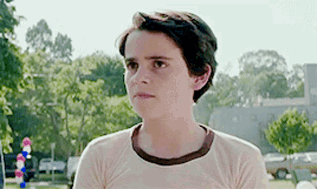 a young boy wearing a tan t-shirt is standing in a park .