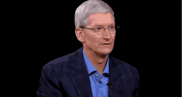 tim cook is wearing glasses and a suit and talking to someone while making a gesture with his hands .