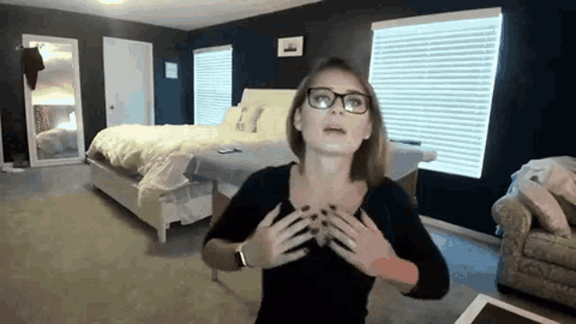 a woman wearing glasses is standing in front of a bed in a bedroom