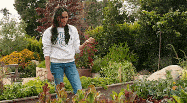 a woman in a white sweatshirt is standing in a garden with lots of plants .