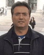 a man wearing glasses and a jacket is standing in front of a building and smiling .