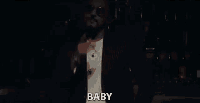 a man wearing sunglasses and a suit is standing in a dark room and says `` baby '' .