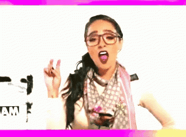 a woman wearing glasses and a scarf is giving a peace sign in front of a sign that says amiquitas