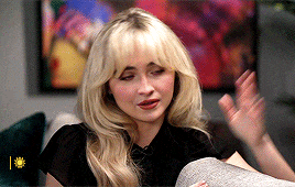 a woman with blonde hair is sitting on a couch in front of a painting and waving her hand .
