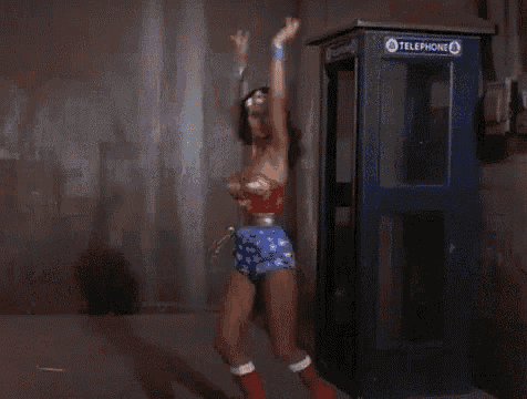 wonder woman is standing in front of a telephone booth with her arms in the air .
