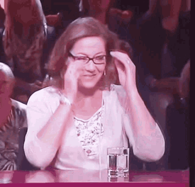 a woman wearing glasses is sitting at a table with a glass of water and clapping her hands .
