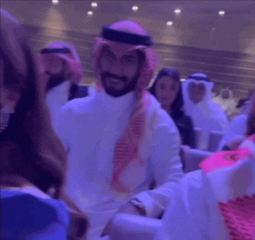 a man wearing a keffiyeh and a hat is sitting in a crowd of people and smiling .