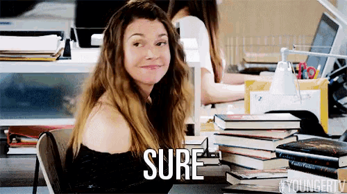 a woman is sitting at a desk with a stack of books and the word sure is on her face .