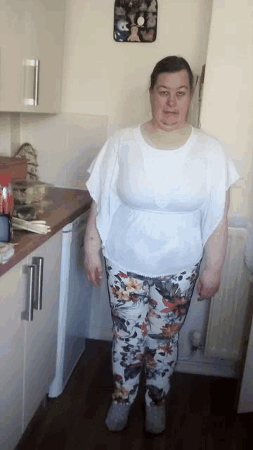 a woman wearing a white top and floral pants stands in a kitchen