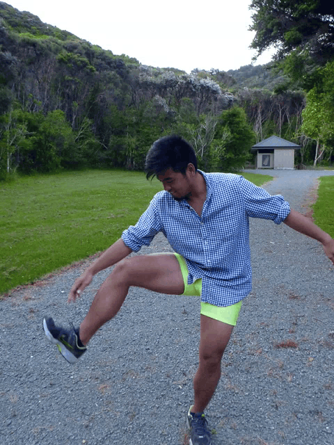 a man in a plaid shirt and neon shorts is standing on one leg on a gravel road