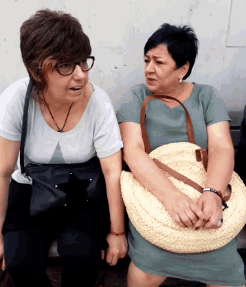 two women are sitting on a bench and one is holding a straw bag