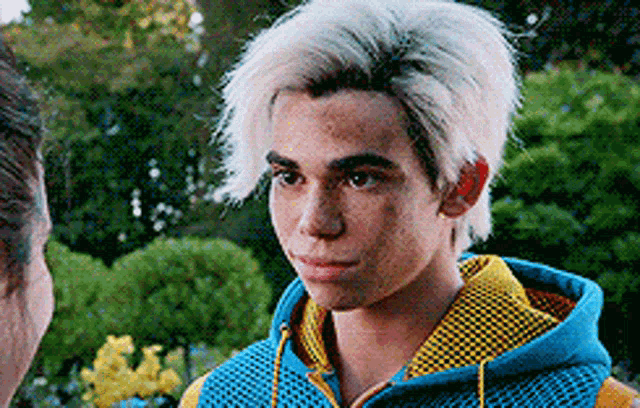 a young man with white hair and freckles is wearing a blue and yellow hoodie and looking at another man .