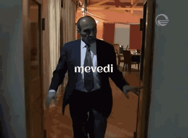 a man in a suit and tie is walking down a hallway with the word mevedi written on the bottom