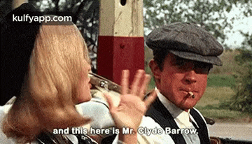a man and a woman are standing next to each other and the man is smoking a cigarette and the woman is waving .