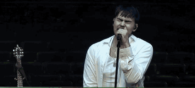 a man in a white shirt singing into a microphone with a guitar in the background