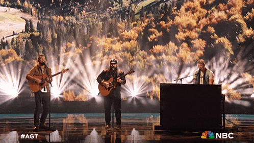 a group of men singing and playing guitars on a stage with a nbc logo in the background