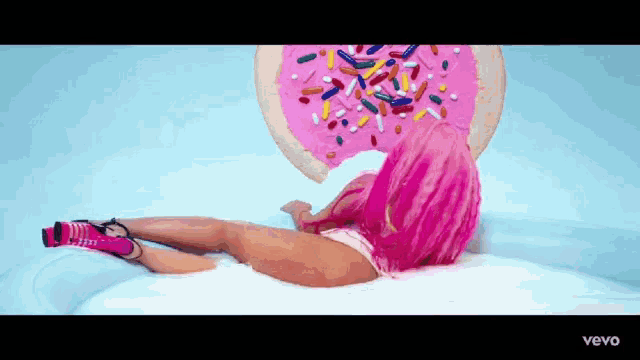 a woman with pink hair is laying on a bed next to a donut with sprinkles .