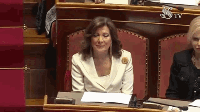 a woman in a white suit sits at a desk in front of a sign that says tv
