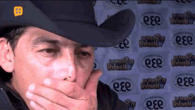 a man wearing a cowboy hat covering his mouth with his hand in front of a wall that says ese