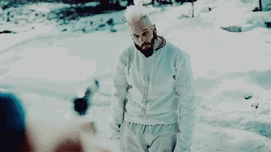 a man with white hair and a beard is standing in the snow with his hands in his pockets