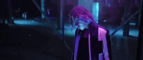 a man with purple hair is standing in a dark room in a neon light .