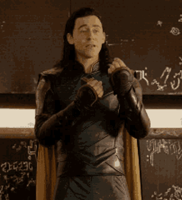 a man in a superhero costume is standing in front of a blackboard and giving a thumbs up .