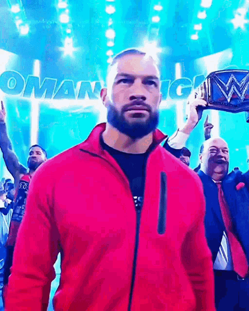 a man in a red jacket stands in front of a sign that says ' roman reigns '