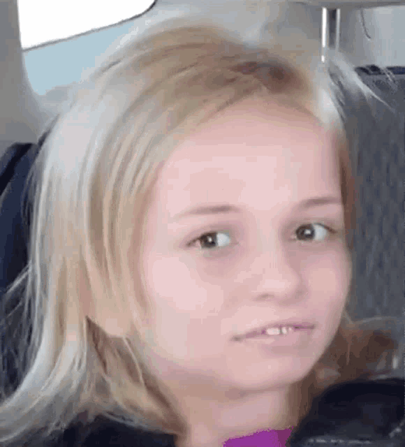 a little girl with blonde hair is sitting in a car making a funny face .