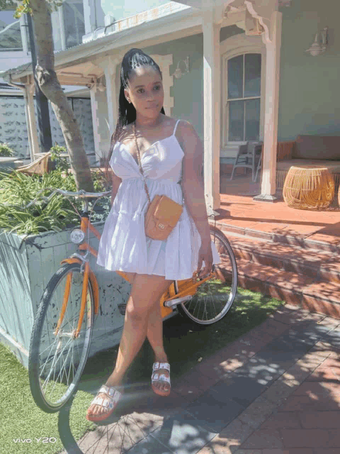 a woman in a white dress is standing next to an orange bicycle taken by a vivo y20