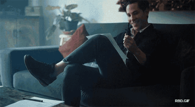 a man is sitting on a couch with his legs crossed and a rbd.gif behind him