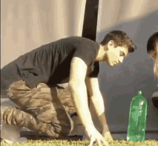 a man is kneeling down in the grass next to a green bottle of water .
