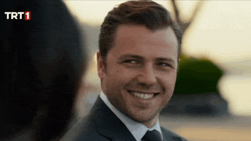 a man in a suit and tie smiles in front of a trt 1 logo