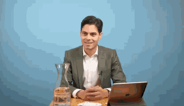 a man in a suit sits at a table with an apple laptop and a pitcher of water