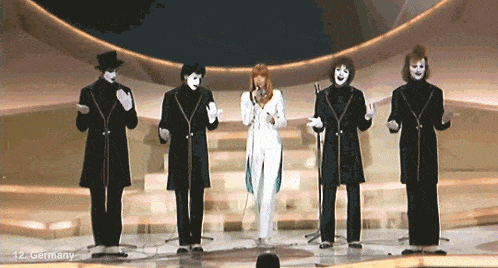 a woman singing into a microphone in front of a group of masked men with the number 12 in the corner