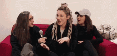 three women are sitting on a red couch talking to each other and holding a microphone .