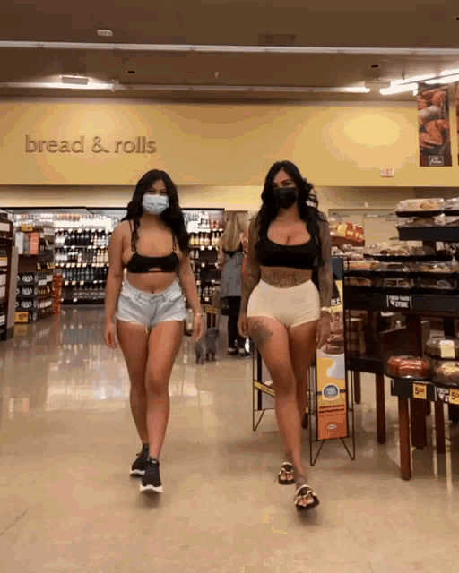 two women wearing face masks are walking through a grocery store aisle that says bread & rolls