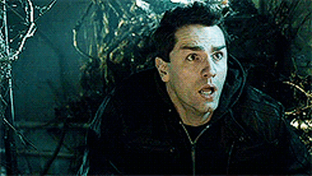 a man in a black jacket is standing in front of a tree looking surprised .