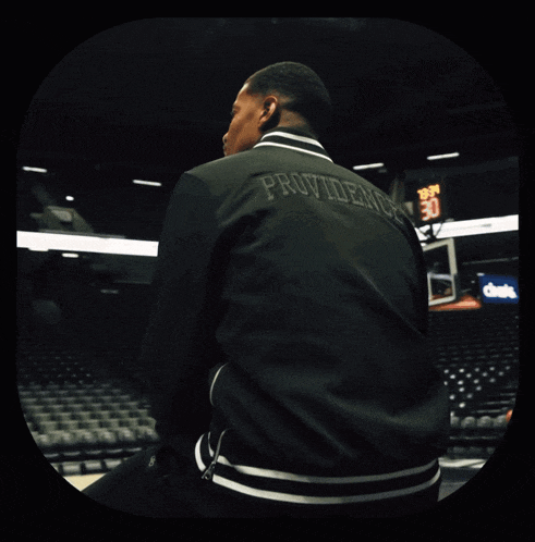 a man wearing a black providence jacket sits in a basketball court