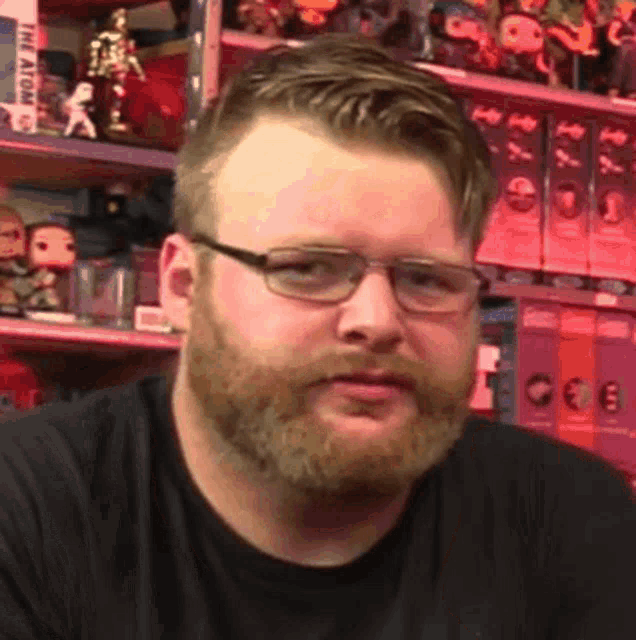 a man with a beard wearing glasses and a black shirt is standing in front of a shelf with toys on it