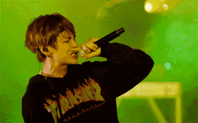 a man wearing a black thrasher sweatshirt sings into a microphone