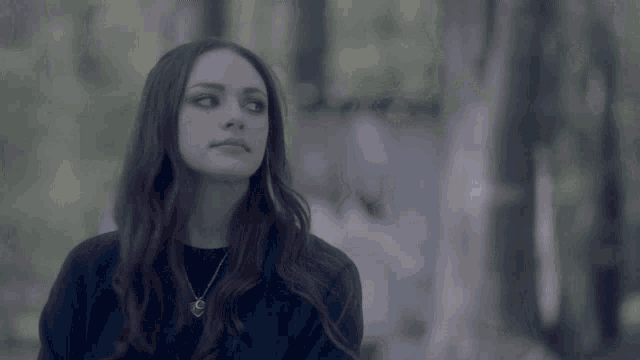 a woman wearing a black sweater and a necklace is standing in a forest
